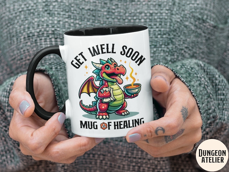May include: White ceramic mug with a black handle. The mug has a cartoon illustration of a red and green dragon holding a bowl of soup. The text on the mug reads "Get Well Soon Mug of Healing".
