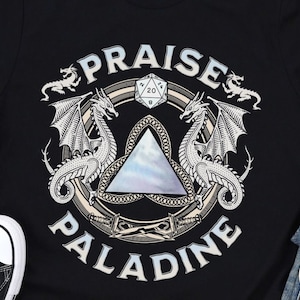 以下が含まれることがあります： ファンタジーデザインの黒いTシャツ。デザインは、円形に配置された「PRAISE PALADINE」の文字が入った中央のエンブレムを挟んで2匹のドラゴンが配置されています。D20サイコロもデザインに含まれています。Dungeon Atelierのロゴが右下にあります。