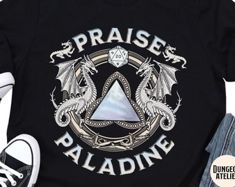 Peut inclure: T-shirt noir avec un motif fantastique. Le motif présente deux dragons encadrant un emblème central avec les mots "PRAISE PALADINE" disposés en cercle. Un dé D20 est également inclus dans le design. Le logo Dungeon Atelier est en bas à droite.