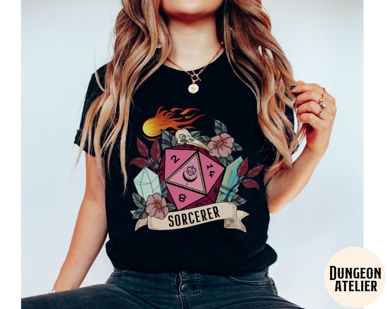 Puede incluir: Camiseta negra con un gr&aacute;fico de dados rosa y blanco, detalles florales y el texto "SORCERER".