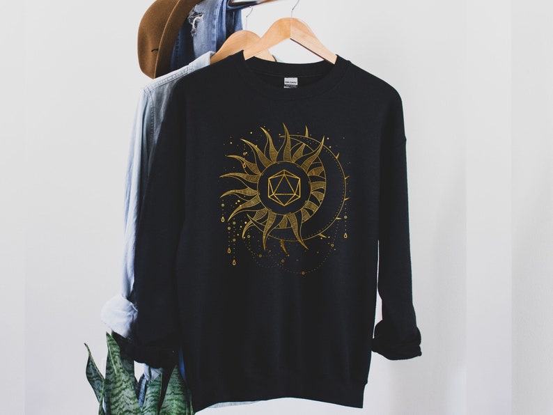 Puede incluir: Sudadera negra con un dise&ntilde;o dorado de sol y luna. El sol presenta una forma geom&eacute;trica en el centro. El dise&ntilde;o incluye puntos y l&iacute;neas doradas. La sudadera est&aacute; colgada en una percha de madera.