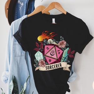 Puede incluir: Camiseta negra con un gr&aacute;fico de dados rosa y rojo, rodeado de flores, cristales y llamas. El dado tiene los n&uacute;meros 2, 8, 14 y 5. El texto "SORCERER" est&aacute; debajo del dado.