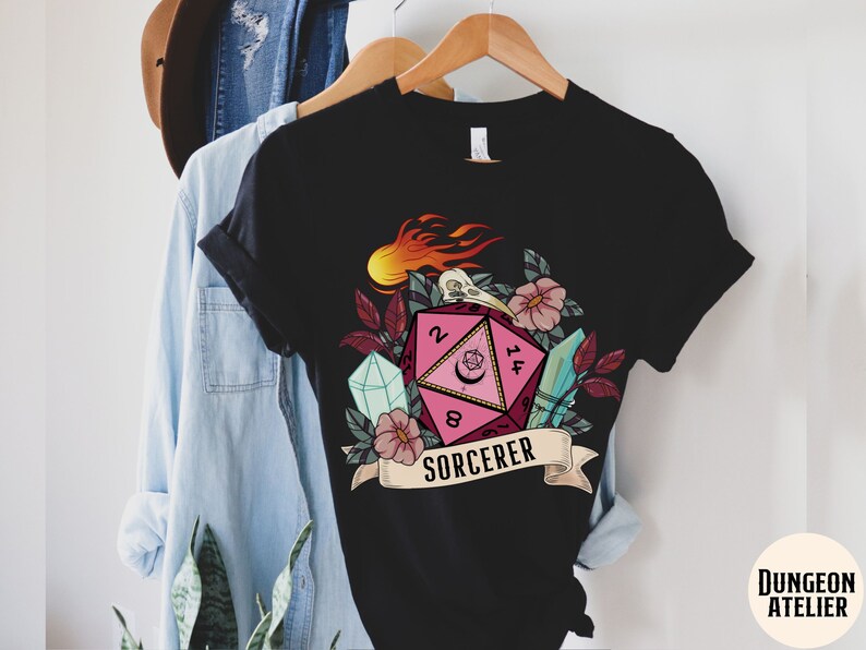 Puede incluir: Camiseta negra con un gr&aacute;fico de dados rosa y rojo, rodeado de flores, cristales y llamas. El dado tiene los n&uacute;meros 2, 8, 14 y 5. El texto "SORCERER" est&aacute; debajo del dado.