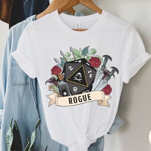 May include: White t-shirt with a black and white illustration of a twenty-sided die, a vial, and daggers. The illustration is surrounded by red roses and green leaves. The text "Rogue" is printed below the illustration.