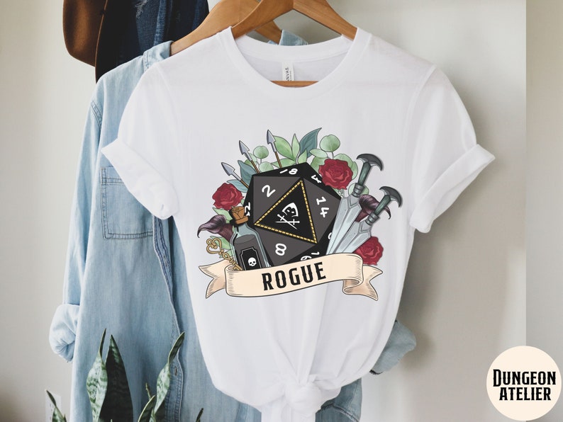 May include: White t-shirt with a black and white illustration of a twenty-sided die, a vial, and daggers. The illustration is surrounded by red roses and green leaves. The text "Rogue" is printed below the illustration.