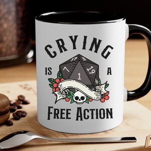 May include: White ceramic mug with a black rim and handle. The mug features a black and white illustration of a twenty-sided die with the text "Crying is a Free Action".