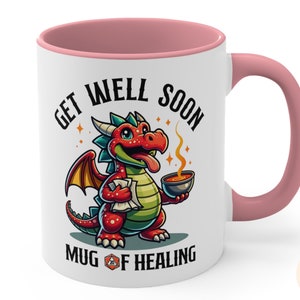 May include: White ceramic mug with a pink rim and handle. The mug features a cartoon red dragon holding a bowl of soup. The text on the mug reads "Get Well Soon Mug of Healing" with a 20-sided die graphic.