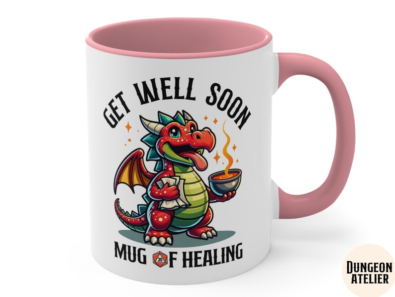 May include: White ceramic mug with a pink rim and handle. The mug features a cartoon red dragon holding a bowl of soup. The text on the mug reads "Get Well Soon Mug of Healing" with a 20-sided die graphic.