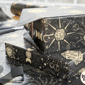 May include: Black and gold wrapping paper with a celestial design featuring dice, moons, stars, and a sun. The paper has the text "Dungeon Atelier" in the bottom right corner.