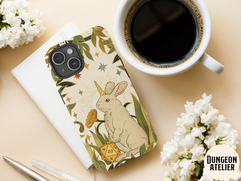 May include: A phone case featuring a whimsical illustration of a unicorn rabbit with a golden horn, surrounded by foliage and a 20-sided die. The case has a cream background with green, yellow, and orange accents. The Dungeon Atelier logo is in the bottom right corner.