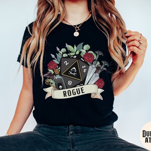May include: Black t-shirt with a graphic of a black twenty-sided die with a skull and crossbones, surrounded by flowers, daggers, and a potion bottle. The text "Rogue" is on a banner below the die.