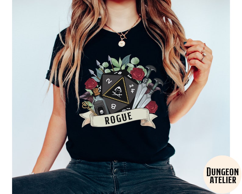 May include: Black t-shirt with a graphic of a black twenty-sided die with a skull and crossbones, surrounded by flowers, daggers, and a potion bottle. The text "Rogue" is on a banner below the die.