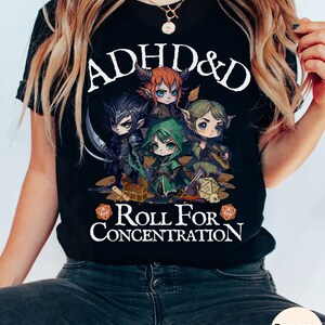 Puede incluir: Camiseta negra con un gr&aacute;fico blanco de cuatro personajes de dibujos animados jugando a Dungeons & Dragons. El texto "ADHD & D Roll For Concentration" est&aacute; impreso en la camiseta.