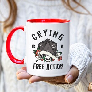 May include: White ceramic mug with a red rim and handle. The mug has a black and white graphic of a twenty-sided die with the text "Crying is a Free Action".