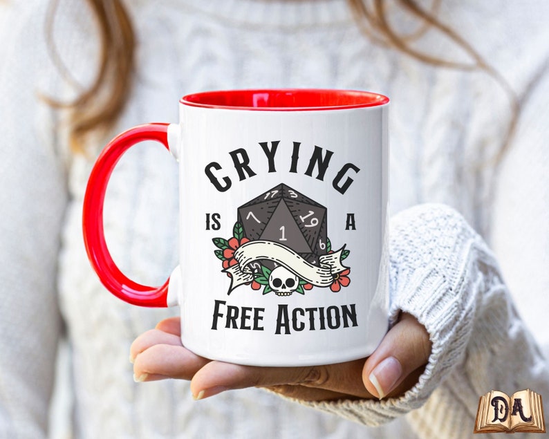 May include: White ceramic mug with a red rim and handle. The mug has a black and white graphic of a twenty-sided die with the text "Crying is a Free Action".
