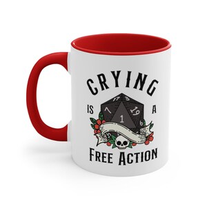 May include: White ceramic mug with a red rim. The mug features a black and white illustration of a twenty-sided die with the text "Crying is a Free Action".