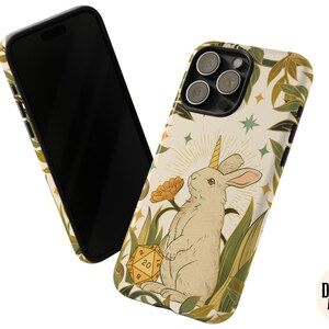 May include: A phone case featuring a whimsical illustration of a rabbit with a unicorn horn, surrounded by foliage and a twenty-sided die. The design is set against a cream background with a black border. The Dungeon Atelier logo is visible.