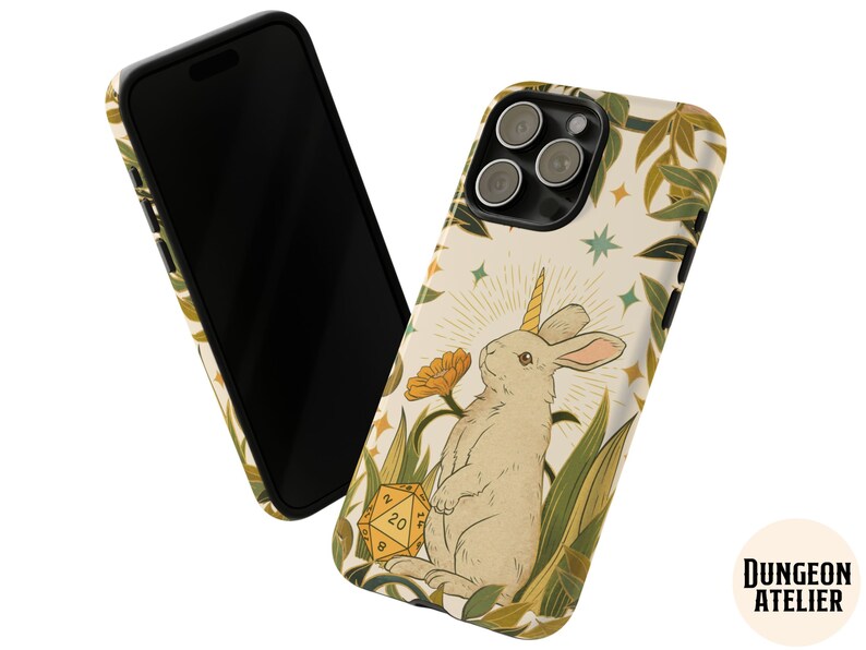 May include: A phone case featuring a whimsical illustration of a rabbit with a unicorn horn, surrounded by foliage and a twenty-sided die. The design is set against a cream background with a black border. The Dungeon Atelier logo is visible.