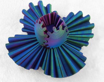 Op de afbeelding: Een cirkelvormig, geplooid sculptuur met een levendig, iriserend verloop van blauwe, paarse en groene tinten. Het midden bestaat uit een kleine, open, tandwielachtige structuur met gekartelde randen, omringd door brede, waaiervormige plooien.