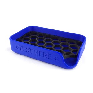 May include: A blue rectangular soap dish with a black honeycomb-patterned insert. The dish has rounded edges and the text "TEXT HERE" is embossed on the side. The soap dish is made of plastic.