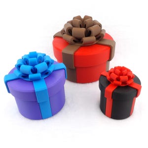 Puede incluir: Tres cajas de regalo con lazos de diferentes colores. La caja más grande es roja con un lazo marrón, la caja mediana es morada con un lazo azul y la caja más pequeña es negra con un lazo rojo.