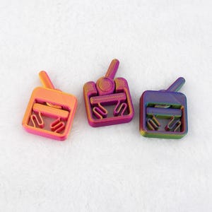 May include: Three colorful, 3D-printed tank-shaped toys. Each toy has a square body with a curved handle and a small antenna. The tanks are in shades of orange, pink, purple, and green, with a rainbow-like effect.