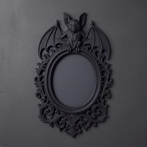 May include: A matte black oval picture frame featuring an ornate, scrollwork border and a sculpted bat figure perched at the top. The frame has a solid black center panel and intricate, symmetrical relief detailing surrounding the entire oval opening.