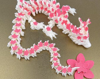 May include: A flexible, articulated dragon figurine constructed from white plastic components. The dragon features a series of small, bright pink flower-shaped accents along its entire length, with a larger pink flower attached to its tail end.