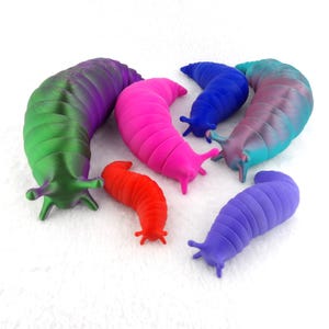 Puede incluir: Un grupo de seis babosas segmentadas, impresas en 3D y de colores vivos. Las babosas son de varios tonos de verde, morado, rosa, azul y rojo. Están dispuestas sobre una superficie blanca y esponjosa.