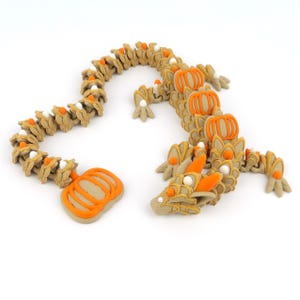 Könnte beinhalten: Gelenkige Drachenfigur in Beige, Orange und Weiß. Der Drachenkörper ist segmentiert, mit Kürbis-Akzenten und einem detaillierten Kopf. Die Figur hat ein Halloween- oder Herbstthema.