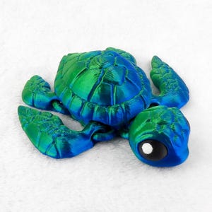 May include: A 3D printed sea turtle figurine with a green and blue iridescent finish. The turtle is lying on its belly with its head turned to the side. The turtle has large, black eyes with white pupils.