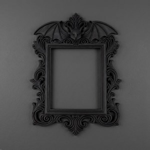 May include: A matte black rectangular picture frame featuring intricate scrollwork and ornate carvings. The top centre of the frame is adorned with a sculpted bat head flanked by two symmetrical, outstretched bat wings, creating a dark aesthetic.
