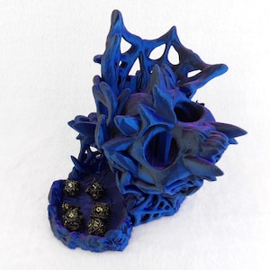 Puede incluir: Una torre de dados con forma de calavera impresa en 3D en azul y morado, con una estructura de telaraña y una bandeja a juego en la base. La torre presenta protuberancias cristalinas afiladas y contiene cinco dados poliédricos negros.