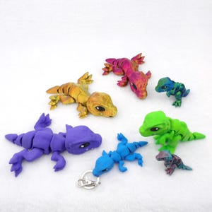Puede incluir: Colección de figuras de lagartos hechas a mano en varios colores. Los lagartos son de color morado, azul, verde, dorado y rosa. Un lagarto azul tiene un llavero. Las figuras tienen detalles.