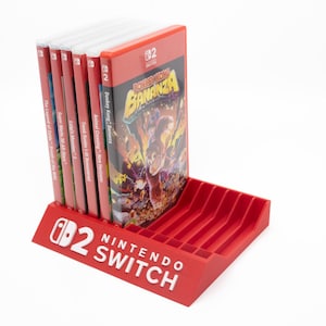 Puede incluir: Un soporte rojo para juegos de Nintendo Switch con varias carcasas de juegos. El soporte tiene ranuras para los cartuchos de juego y el logotipo de Nintendo Switch. Las carcasas de los juegos son rojas y presentan títulos como "Donkey Kong Country: Tropical Freeze".