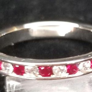 May include: A silver ring with alternating red and clear gemstones set around the band. The gemstones are evenly spaced, creating a continuous pattern. The ring is displayed against a dark background, highlighting its sparkle.