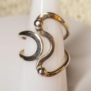 May include: A silver-toned ring with a unique, abstract design. The ring features curved, flowing lines and small spherical accents. The ring is displayed on a white, cylindrical form, showcasing its shape and style. The ring's design is modern and eye-catching.