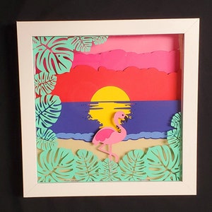 Pode incluir: Uma moldura branca com um recorte de papel de um flamingo rosa em pé numa praia com um sol amarelo e folhas de palmeira verdes.