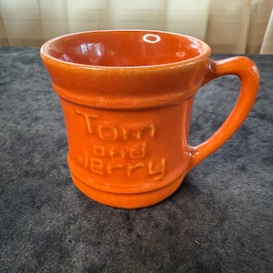 以下が含まれることがあります： オレンジ色の取っ手付きセラミックマグ。マグカップの前面には「Tom and Jerry」のエンボス加工が施されています。マグカップは口が広く、底が丸みを帯びています。マグカップは単色のオレンジ色です。