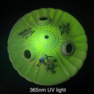 Puede incluir: Un objeto cerámico redondo de color verde claro con tres aberturas cilíndricas y un agujero central. Se ven diseños florales y un patrón central en relieve. El objeto brilla intensamente bajo la luz UV de 365 nm.