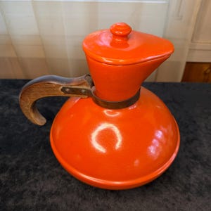 May include: A vintage orange ceramic coffee pot with a wooden handle and lid. The pot has a round base and a spout. The handle is attached with metal hardware. The lid has a small knob. The coffee pot is a retro kitchen item.