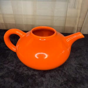Könnte beinhalten: Eine leuchtend orangefarbene Keramik-Teekanne mit rundem Körper, gebogenem Griff und Ausguss. Die Teekanne hat eine glänzende Oberfläche und ist zum Aufbrühen und Servieren von Tee konzipiert. Der Griff ist groß und leicht zu greifen.