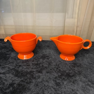 Könnte beinhalten: Eine leuchtend orangefarbene Zuckerdose aus Keramik mit zwei Griffen und ein passender Sahnegießer. Beide Teile haben einen Sockel und horizontale geriffelte Details. Der Sahnegießer hat einen Ausguss und einen einzelnen Griff. Das Set wird auf einer dunklen Oberfläche präsentiert.
