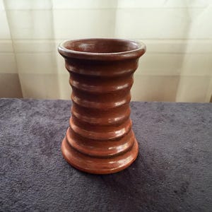 Może przedstawiać: Brązowy ceramiczny wazon o cylindrycznym kształcie z serią poziomych, wypukłych prążków biegnących wzdłuż zewnętrznej strony. Wazon posiada szeroki, rozszerzający się ku górze kołnierz oraz nieco szerszą podstawę, z gładkim, błyszczącym wykończeniem.
