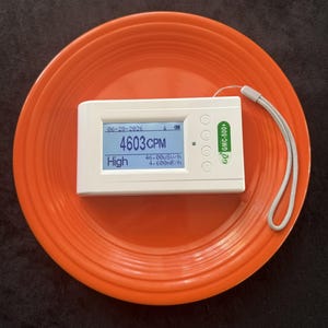 May include: A white GQ GMC-500+ Geiger counter with a digital display showing 4603 CPM, 46.00uSv/h, and 4.600mR/h, dated 06-28-2026, featuring a grey wrist strap, resting on a bright orange circular plate with concentric ridged detailing.