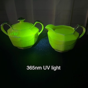 以下が含まれることがあります： UVライトの下で光る緑色のティーセット。セットには、蓋付きのティーポットとクリーマーが含まれており、どちらもリブ付きの側面と濃いアクセントが付いています。「365nm UV light」というテキストが下に表示されています。