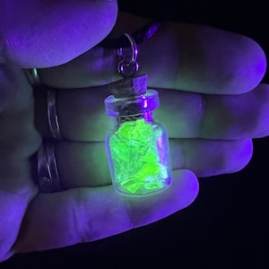 May include: A small glass vial pendant filled with glowing neon green fragments. The vial has a cork stopper and is suspended from a silver-colored bail on a black cord. The background is dark.
