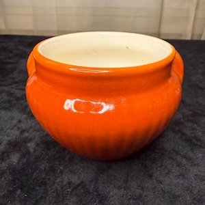 May include: A small, orange ceramic pot with a glossy finish. The pot has a rounded shape with two small handles and a cream-colored interior. The pot is likely used for cooking or serving.