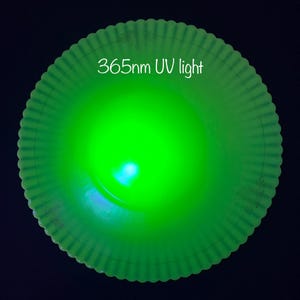 May include: A glowing green, round object with a scalloped edge, illuminated by 365nm UV light. The object emits a bright, vibrant green color, with the text "365nm UV light" in white.