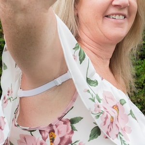 May include: A white floral print top with pink roses and green leaves. The top has a white adjustable strap. The background is a green, out-of-focus natural setting.